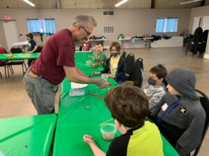 A master gardener volunteer is standing at a table teaching five young boys about gardening.